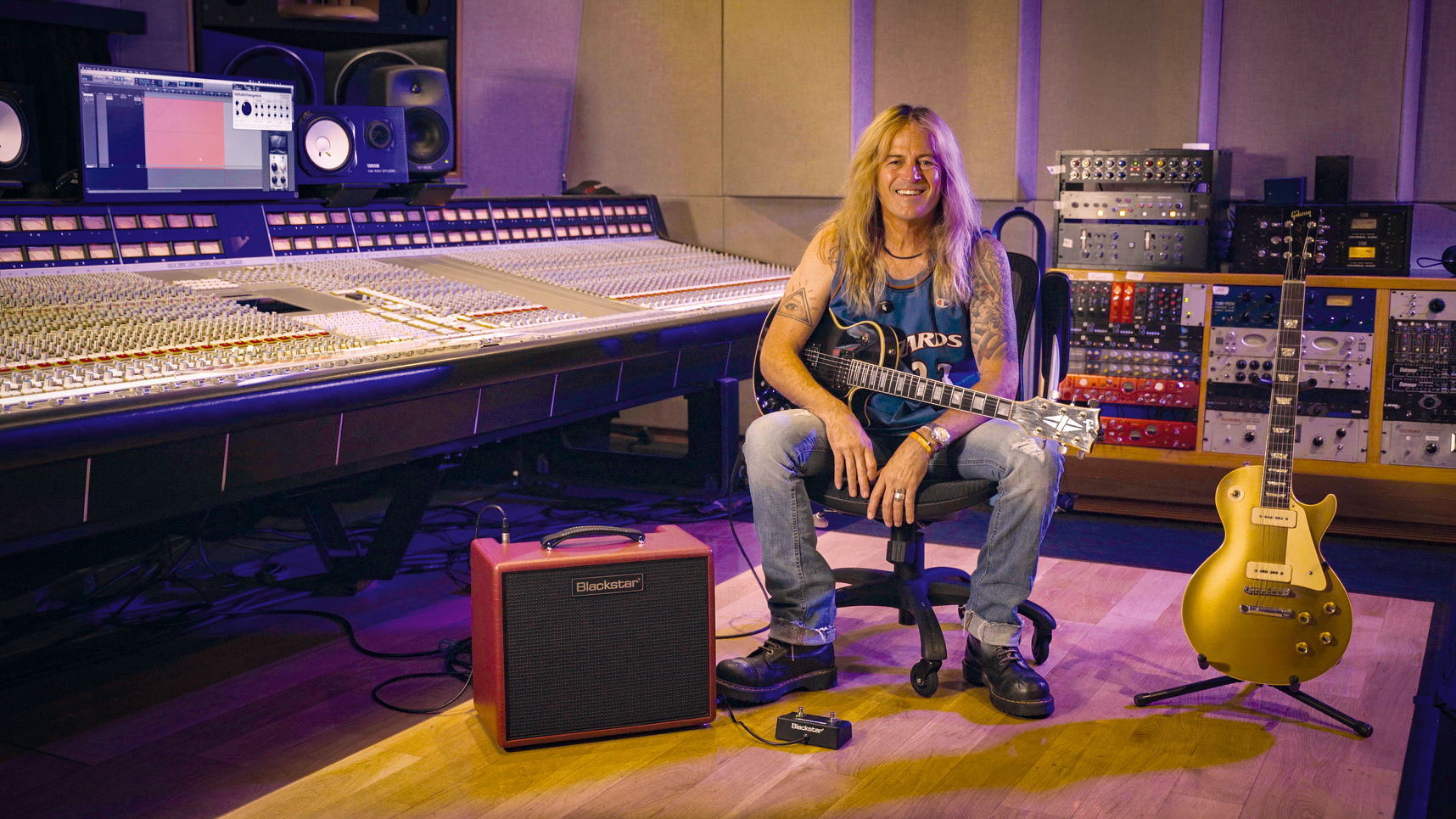 A woman sits in a recording studio chair holding a black electric guitar, with a red Marshall amplifier in the foreground and a gold electric guitar on a stand to her right. She is in front of a large mixing console and audio equipment.