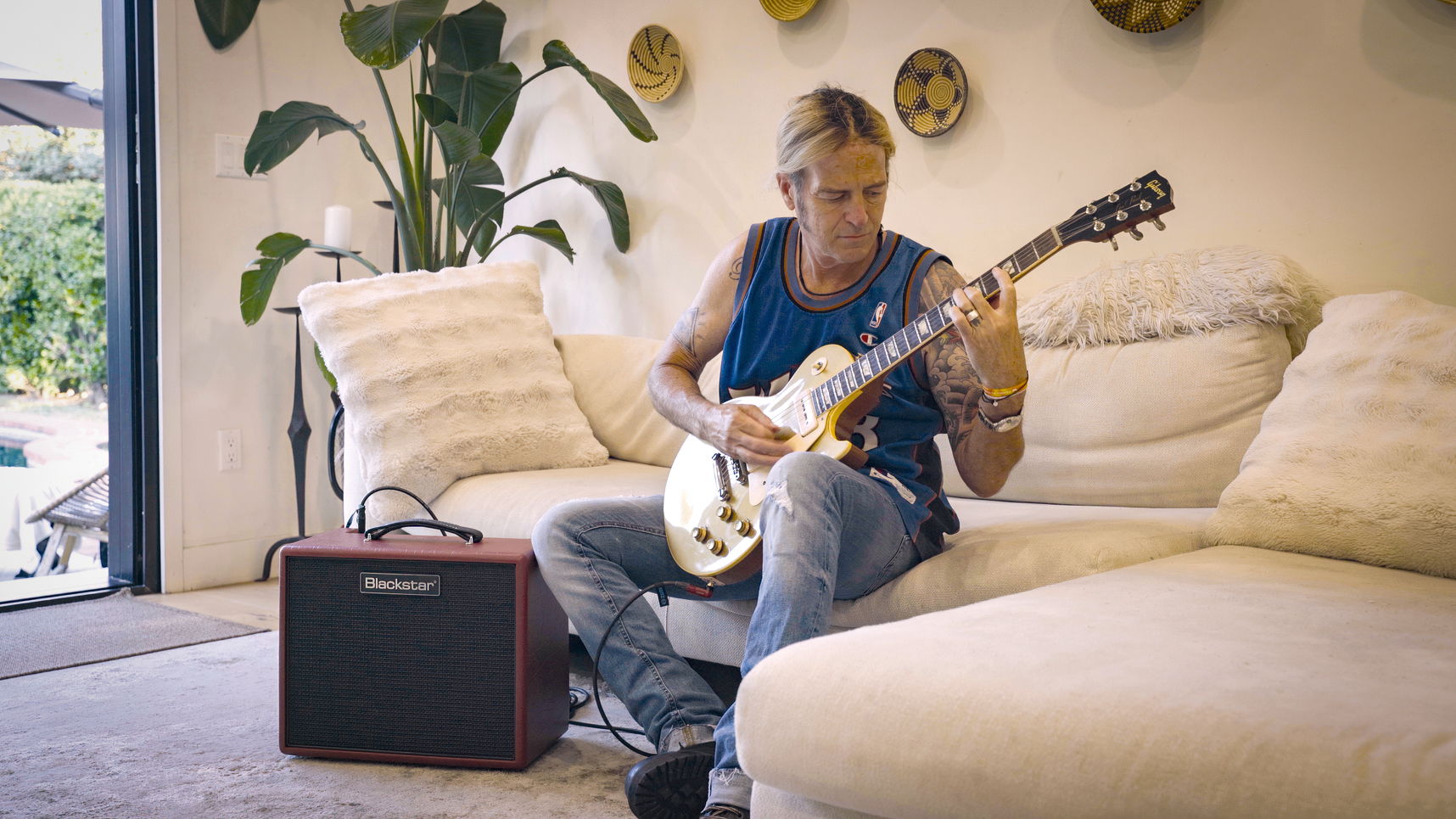 A man sits on a cream sectional sofa playing a gold electric guitar, with a Marshall speaker and cable on the floor next to him in a living room setting.