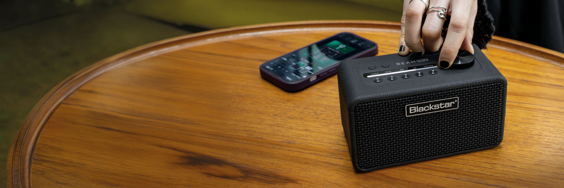 Blackstar BEAM Mini amplifier bluetooth speaker on a wooden table with a hand adjusting the speed dial and a mobile phone app displayed nearby